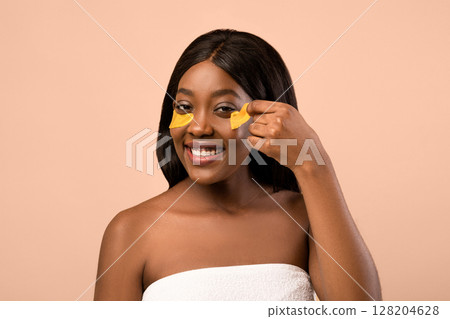Closeup of smiling beautiful young black lady removing golden hydrogel eye patches over beige studio background, copy space. Anti-aging, against lines cosmetic tools, wrinkles eye care concept 128204628