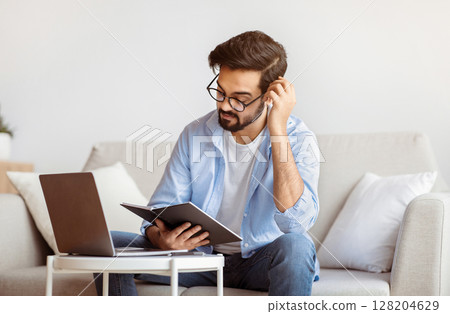 Young Arab Freelancer Guy Checking Working Schedule In Planner Notepad At Home. Millennial Eastern Man Sitting At Table With Laptop Computer, Enjoying Remote Work And Distance Job Opportunities 128204629