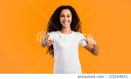 I'm Cool. Cheerful Young Woman In White T-Shirt Gesturing Thumbs Up Pointing Finger At Herself Standing On Yellow Background In Studio, Smiling To Camera. Choose Me Concept. Panorama 128204631