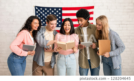New study, learning and sharing knowledge. Surprised young multiracial students with notebooks and gadgets look in book in hands of asian woman, on white brick wall background with big USA flag New study, learning and sharing knowledge. Surprised young multiracial students with notebooks and gadgets look in book in hands of asian woman, on white brick wall background with big USA flag 128204645