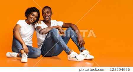 Attractive black couple sitting on floor together, posing at studio, orange background, copy space. Smiling african american young woman leaning on her happy boyfriend, spending time together Attractive black couple sitting on floor together, posing at studio, orange background, copy space. Smiling african american young woman leaning on her happy boyfriend, spending time together 128204656