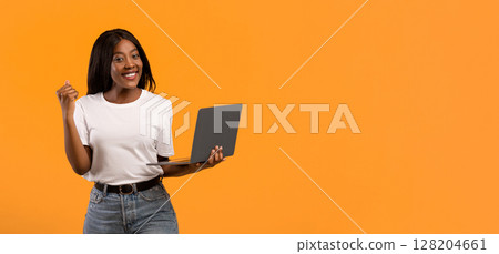 Smiling young black woman in white t-shirt holding laptop and raising fist up over orange studio background, catch nice discounts for online shopping or celebrating success in gambling, copy space Smiling young black woman in white t-shirt holding laptop and raising fist up over orange studio background, catch nice discounts for online shopping or celebrating success in gambling, copy space 128204661