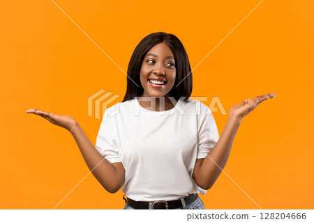 Confused black lady raising both hands up as holding something invisible on orange studio background, copy space. Smiling african american young woman showing or recommending something 128204666