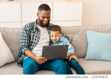 African Daddy And Son Watching Cartoons Online On Laptop Computer Sitting On Sofa Together At Home. Weekend With Father. Family Lifestyle And Gadgets Concept 128204673