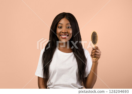 Attractive young black woman holding wooden hair brush and smiling over beige studio background. Happy african american lady with beautiful long hair enjoying her new wooden hair brush 128204693