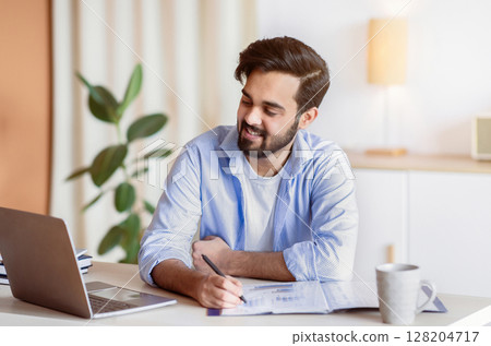 Successful Eastern Entrepreneur Working With Documents And Laptop At Home Office, Smiling Arab Businessman Looking At Computer Screen, Checking Financial Reports And Taking Notes, Free Space 128204717