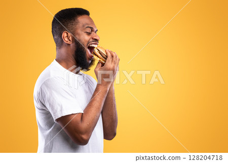Black Millennial Guy Biting Burger Eating Junk Food Over Yellow Studio Background. Side View, Blank Space. African American Man Enjoying Tasty Cheeseburger. Overeating, Unhealthy Nutrition Habit 128204718