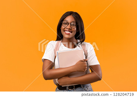 Smiling young african american lady student with backpack, books and notebooks posing over orange studio background, wearing glasses, copy space. Education, college, university concept 128204719