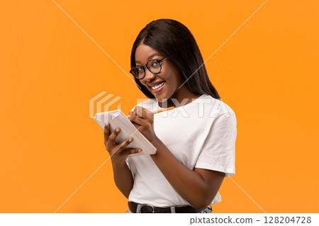 Clever young black woman in glasses smiling and taking notes, holding notepad and pen, orange studio background, copy space. Cheerful intelligent african american lady planning day Clever young black woman in glasses smiling and taking notes, holding notepad and pen, orange studio background, copy space. Cheerful intelligent african american lady planning day 128204728