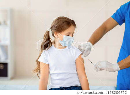 Black doctor making coronavirus vaccine injection to little girl in protective mask at hospital. Child being immunized against covid-19 at health centre. Global vaccination campaign concept 128204732