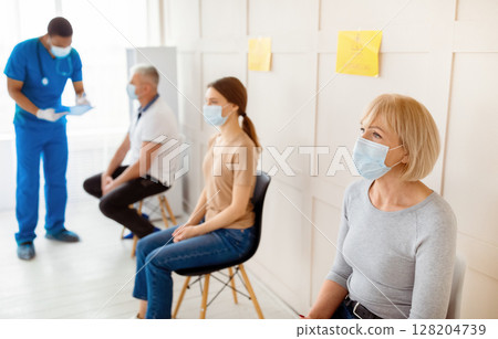 Mature woman and other patients in face masks waiting in queue for vaccination against covid-19 at clinic, communicating to doctor, taking part in anti coronavirus campaign, copy space 128204739