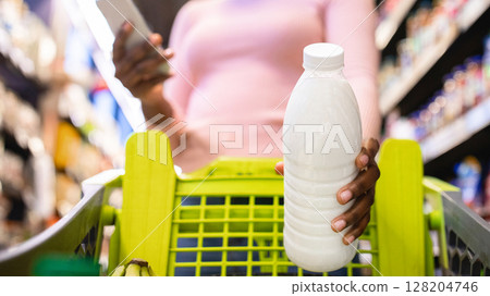 Young black woman checking her shopping list in smartphone app, putting bottle of milk into her cart at supermarket, selective focus. Low angle view. African American lady buying groceries at mall Young black woman checking her shopping list in smartphone app, putting bottle of milk into her cart at supermarket, selective focus. Low angle view. African American lady buying groceries at mall 128204746