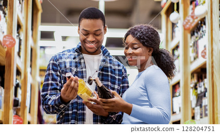 Cheerful black couple holding wine bottles, selecting alcohol beverages at modern supermarket. Lovely young African American family buying drinks at liquor store or huge mall Cheerful black couple holding wine bottles, selecting alcohol beverages at modern supermarket. Lovely young African American family buying drinks at liquor store or huge mall 128204751