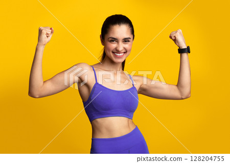 Winner. Portrait of excited young woman in sportswear celebrating triumph, raising clenched fists in the air isolated over orange yellow studio background. Happy smiling lady posing 128204755