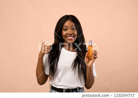 Pretty young black woman with long beautiful hair holding bottle with hair product, serum or oil, beige studio background, copy space. Hair care, healthy and beautiful hair concept 128204757
