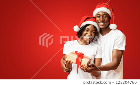 Happy black couple celebrating New Year together, wearing Santa hats, embracing, holding gift box and smiling at camera over orange studio background, copy space. Winter holidays concept 128204760