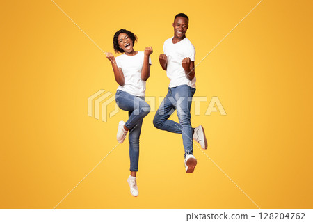 Loving black couple expressing positive emotions, raising fists together and jumping, orange studio background, full length shot. Excited african american young man and woman posing together 128204762