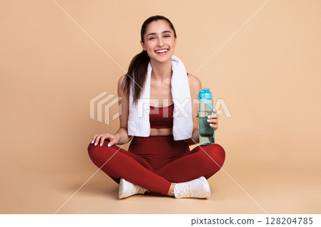 Healthy beautiful smiling young woman in sportswear and white towel on neck sitting on the floor and holding bottle with fresh water, looking at camera isolated over pink studio background, banner Healthy beautiful smiling young woman in sportswear and white towel on neck sitting on the floor and holding bottle with fresh water, looking at camera isolated over pink studio background, banner 128204785