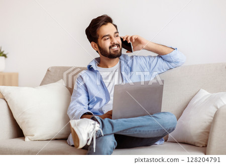 Remote Work Concept. Handsome Arab Male Freelancer Using Laptop And Talking On Cellphone At Home, Sitting With Computer On Couch In Living Room, Enjoying Distant Job Opportunities, Copy Space 128204791