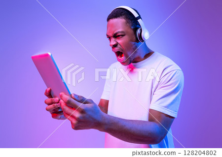 Online Gaming. Angry African Man In Wireless Headphones Using Digital Tablet, Looking At Glowing Screen And Shouting With Rage, Emotionally Reacting To Game Loss, Standing In Neon Purple Light 128204802