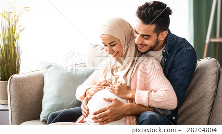 Happy islamic couple expecting baby, hugging on couch at home, cheerful muslim spouses relaxing together on sofa in living room, tenderly touching pregnant belly and smiling, copy space Happy islamic couple expecting baby, hugging on couch at home, cheerful muslim spouses relaxing together on sofa in living room, tenderly touching pregnant belly and smiling, copy space 128204804