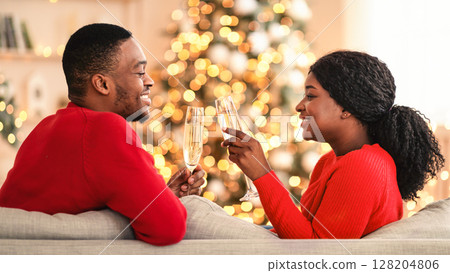Christmas, New Year and comfort of home. Happy young african american couple in love clink glasses with champagne, sit on sofa with tree with garlands in modern living room interior, free space 128204806