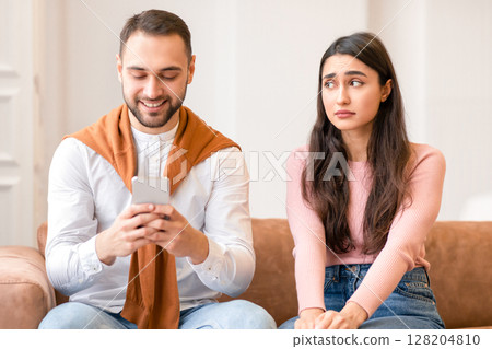 Sad Arab Wife Looking At Husband While He Texting On Cellphone Ignoring Her Sitting On Sofa At Home. Woman Suspecting Infidelity In Marriage. Family And Relationship Problem Concept 128204810
