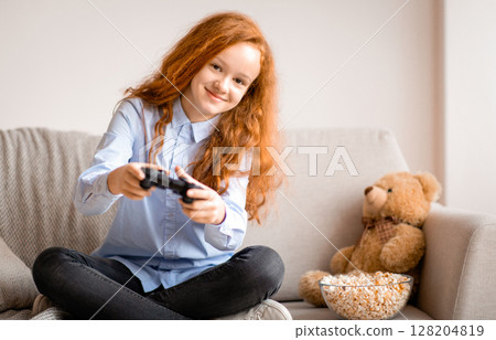 Young Gamer. Portrait of smiling teenage red-haired girl single playing a video game with a black controller, holding joystick, looking at camera, sitting on the couch in living room at home Young Gamer. Portrait of smiling teenage red-haired girl single playing a video game with a black controller, holding joystick, looking at camera, sitting on the couch in living room at home 128204819