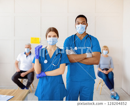 Male and female doctors wearing uniforms and face masks, holding syringe with coronavirus vaccine at clinic, patients waiting for covid-19 immunization at background. Medical help concept Male and female doctors wearing uniforms and face masks, holding syringe with coronavirus vaccine at clinic, patients waiting for covid-19 immunization at background. Medical help concept 128204852