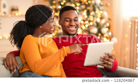Young couple choosing film, resting together at cozy winter evening. Happy millennial african american lady points finger at tablet and shows to guy something interesting in interior with garlands Young couple choosing film, resting together at cozy winter evening. Happy millennial african american lady points finger at tablet and shows to guy something interesting in interior with garlands 128204867