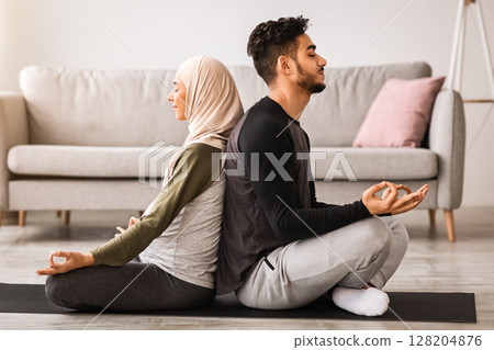 Yoga for Pregnancy and Childbirth. Expecting islamic spouses meditating together at home, pregnant muslim woman in hijab sitting back to back with her husband, practicing lotus pose, side view 128204876