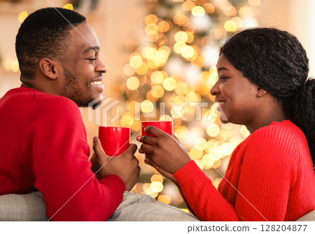 Romance, free time and celebration of covid-19 quarantine. Young happy african american couple with cups of hot drink, sit on sofa in living room interior with Christmas tree with glowing garlands 128204877