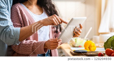 Online Recipe. Cropped shot of young couple using digital tablet in kitchen while cooking food together, unrecognizable spouses checking ingredients in internet while preparing healthy lunch, closeup 128204930