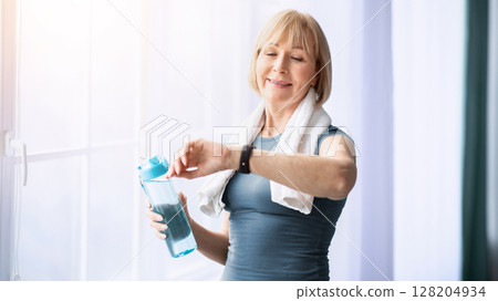 Portrait of sporty senior woman with bottle of water checking her watch or fitness tracker indoors. Athletic mature lady staying hydrated during sports training, leading healthy lifestyle 128204934