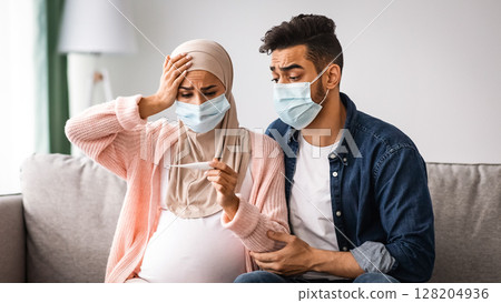 Coronavirus During Pregnancy. Worried Pregnant Muslim Woman Wearing Protective Medical Mask Looking At Thermometer While Sitting Next To Her Husband On Couch In Living Room, Feeling Unwell At Home Coronavirus During Pregnancy. Worried Pregnant Muslim Woman Wearing Protective Medical Mask Looking At Thermometer While Sitting Next To Her Husband On Couch In Living Room, Feeling Unwell At Home 128204936