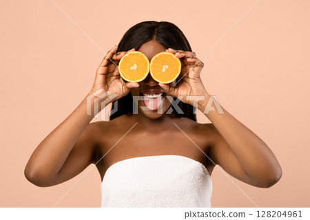 Playful young black woman wrapped in towel covering eyes with halves of orange and smiling, anti-aging procedures, vitamins and healthcare concept, beige studio background, copy space 128204961