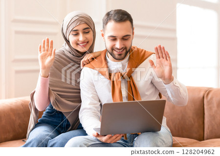 Muslim Family Couple Making Video Call Using Laptop Sitting On Sofa At Home. Cheerful Arab Spouses Video Calling Remote Friend Waving Hand To Computer Webcam. Distant Communication Concept 128204962