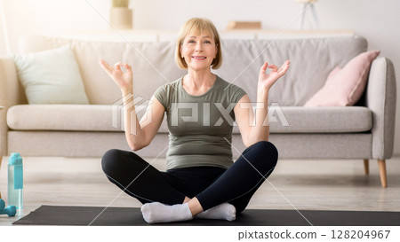 Balanced senior woman sitting in lotus pose and making gyan mudra with her hands, meditating on sports mat at home. Peaceful mature lady practicing yoga and mindfulness, copy space 128204967