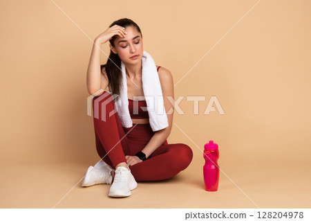 Rest And Relax Concept. Portrait of sporty young woman with white towel on neck sitting on the floor with bottle of water at studio after exercising, taking break. Wellbeing And Training 128204978