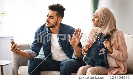 Gadget Addiction Concept. Indifferent arab man using smartphone at home, ignoring his pregnant wife. Upset Expectant Muslim Lady In Hijab Asking Attention From Careless Husband, Free Space 128204981