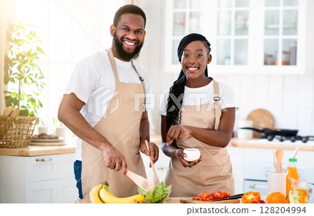Happy Millennial African American Couple Wearing Aprons Cooking Healthy Lunch Together In Kitchen, Cheerful Black Spouses Enjoying Preparing Food At Home, Looking At Camera And Smiling, Free Space Happy Millennial African American Couple Wearing Aprons Cooking Healthy Lunch Together In Kitchen, Cheerful Black Spouses Enjoying Preparing Food At Home, Looking At Camera And Smiling, Free Space 128204994
