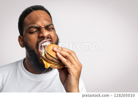 African Man Biting Burger Enjoying Unhealthy Tasty Junk Food Standing In Studio Over White Background. Overeating, Fast Food And Male Nutrition Concept. Funny Hungry Black Guy Eats Cheeseburger 128205010