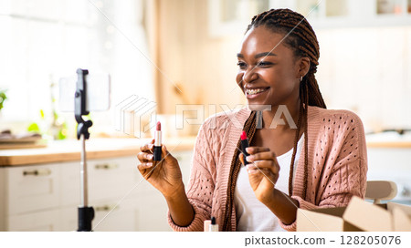 Joyful Young Black Beauty Blogger Lady Recording Makeup Review Video In Kitchen At Home, Happy African American Influencer Woman Holding Two Lipsticks, Using Smartphone On Tripod For Creating Content Joyful Young Black Beauty Blogger Lady Recording Makeup Review Video In Kitchen At Home, Happy African American Influencer Woman Holding Two Lipsticks, Using Smartphone On Tripod For Creating Content 128205076
