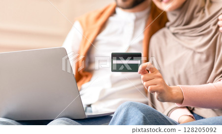Online Shopping. Muslim Couple Using Credit Card And Laptop Computer Buying Things In Internet Shop Sitting On Sofa At Home. Sales And Discounts Concept. Selective Focus, Shallow Depth, Cropped 128205092