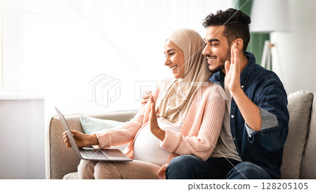 Cheerful Pregnant Muslim Couple Using Laptop At Home For Video Call, Happy Expectant Middle Eastern Spouses Talking To Family Or Friends Via Web Conference, Waving Hands At Computer Camera, Side View 128205105