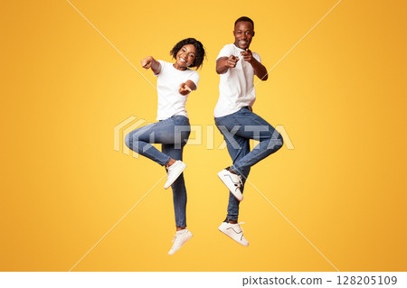 Emotional black couple attractive young man and woman wearing jeans and white t-shirts posing on orange studio background, jumping in the air, pointing fingers at camera. Millennials lifestyle concept 128205109