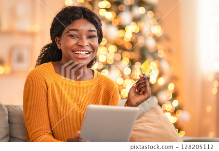 Online shopping at home during covid-19 self-isolation. Millennial african american laughing lady holds tablet and credit card for shopping in living room interior with Xmas tree and glowing garlands 128205112