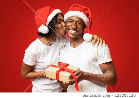 Happy black woman giving her boyfriend Christmas gift on orange studio background, copy space. Cheerful african american family in Santa hats celebrating New Year together, lady kissing man 128205120