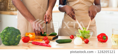 Unrecognizable black couple in aprons preparing vegetable salad together in kitchen. Young african american family cooking healthy dinner together, enjoying making healthy food at come, cropped image 128205121