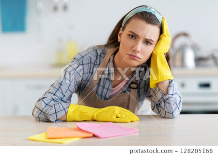 Unhappy young woman tuired from housework at kitchen, needing break from domestic duties. Overworked lady wearing rubber gloves, doing house chores, wiping table with rag Unhappy young woman tuired from housework at kitchen, needing break from domestic duties. Overworked lady wearing rubber gloves, doing house chores, wiping table with rag 128205168